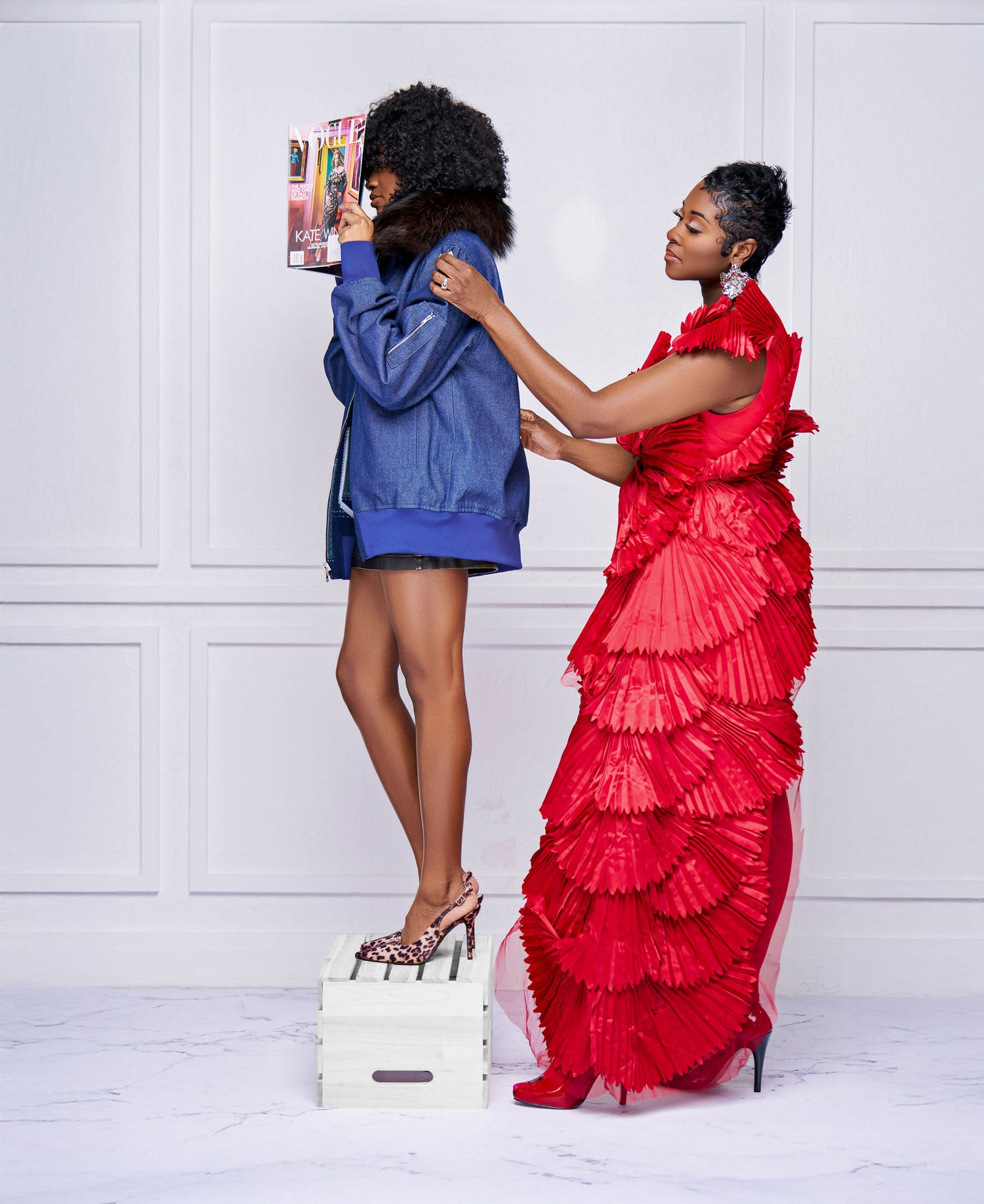 Woman in a red ruffled dress measuring another woman's height against a white paneled wall.
